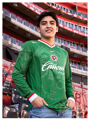 Gilberto Mora posando en gradas del estadio Caliente con jersey verde manga larga Edici&oacute;n Especial Reebok x Club Tijuana, escudo Xoloitzcuintles en pecho, logo Reebok visible, fondo de butacas rojas y negras del estadio, colecci&oacute;n oficial Reebok.