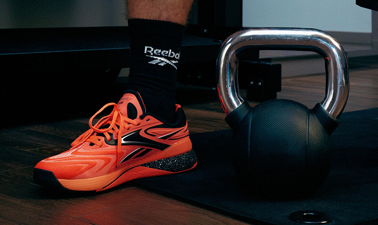 Imagen carrusel Reebok Nano X5 Edge, tenis crossfit naranja ne&oacute;n con detalles negros y plateados junto a kettlebell met&aacute;lico sobre colchoneta negra, calcetines negros con logo Reebok bordado, suela con acabado texturizado, fondo de gimnasio con iluminaci&oacute;n tenue