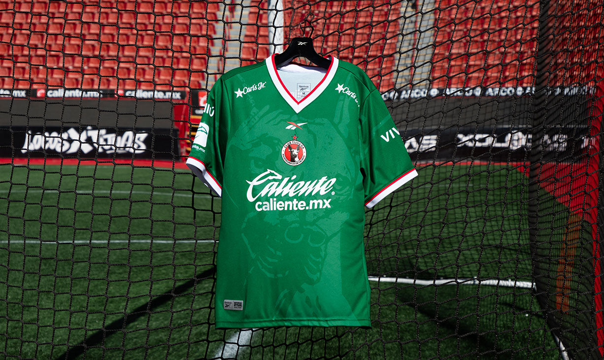Carrusel Reebok x Club Tijuana con jersey verde colgado en porter&iacute;a del estadio, escudo Xoloitzcuintles, patrocinador Caliente, gradas rojas de fondo.
