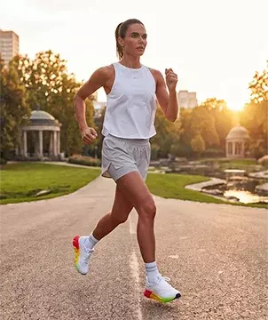 Look de running Reebok SoleMate para mujer, modelo corriendo en carretera al atardecer, tank top blanco Reebok, short gris, calcetines blancos y tenis de running blancos Reebok con suela multicolor arco&iacute;ris, parque y edificios al fondo con luz c&aacute;lida. Ropa deportiva Reebok para mujer, tenis de running blancos SoleMate y outfit para correr.
