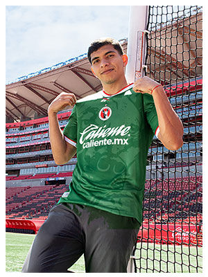 Modelo masculino sosteniendo el cuello del jersey verde manga corta Reebok x Club Tijuana frente a la porter&iacute;a del estadio Caliente, escudo Xoloitzcuintles y logo Reebok visibles, fondo de butacas rojas y estructura met&aacute;lica.