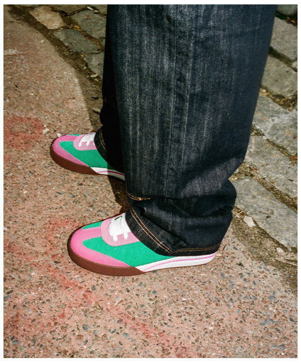Modelo con jeans oscuros calzando tenis skate Reebok x IceCream Board Flip en combinaci&oacute;n verde rosa y blanco con suela caf&eacute;, sobre pavimento texturizado con adoquines grises, estilo urbano diurno.