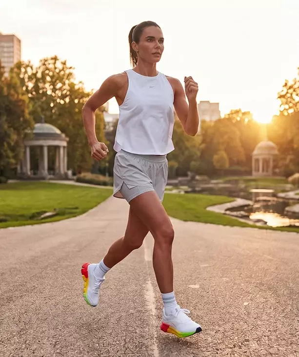 Look de running Reebok SoleMate para mujer, modelo corriendo en carretera al atardecer, tank top blanco Reebok, short gris, calcetines blancos y tenis de running blancos Reebok con suela multicolor arco&iacute;ris, parque y edificios al fondo con luz c&aacute;lida. Ropa deportiva Reebok para mujer, tenis de running blancos SoleMate y outfit para correr.