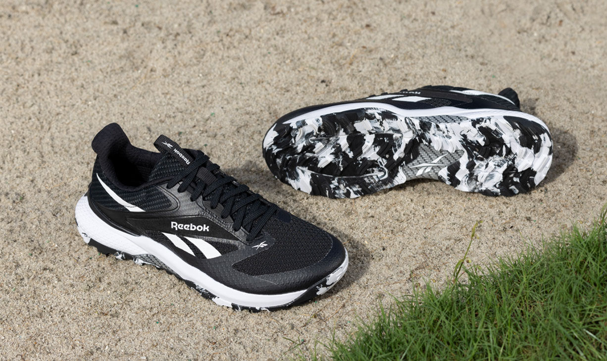 Carrusel Reebok Nano Golf con par de tenis negro sobre arena y pasto, suela con patr&oacute;n camuflaje blanco y negro, logo Reebok vector.