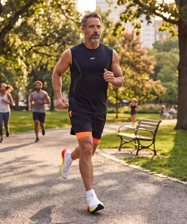 Look de running Reebok SoleMate para hombre, modelo corriendo en un parque arbolado, tank top negro Reebok, short negro con short de compresi&oacute;n naranja, calcetines blancos y tenis de running blancos Reebok con suela multicolor arco&iacute;ris, otros corredores al fondo. Ropa deportiva Reebok para hombre, tenis de running blancos SoleMate y outfit para correr.