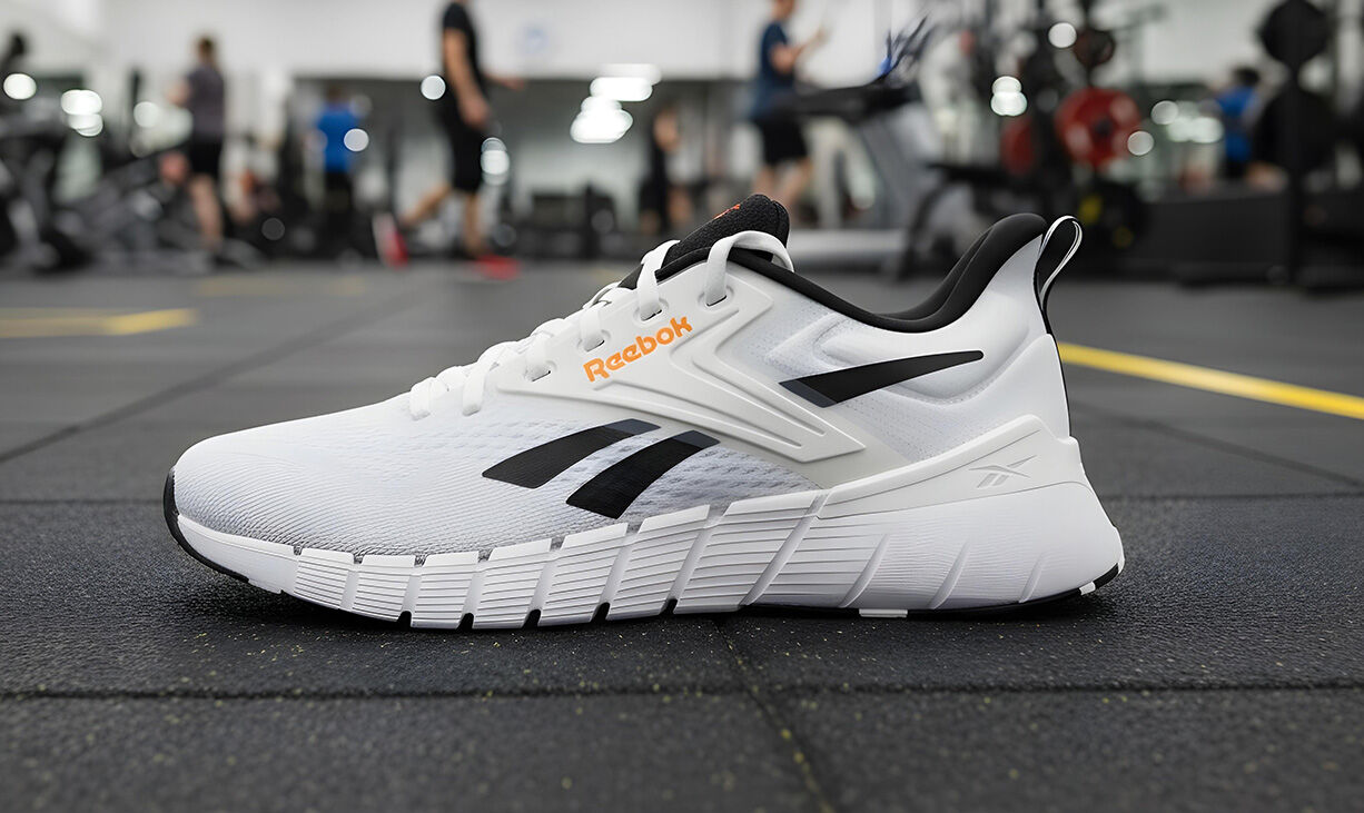 Tenis Reebok Nano Gym blanco con acentos amarillo ne&oacute;n y negro, posicionado en piso de gimnasio con equipo de entrenamiento desenfocado al fondo, dise&ntilde;o t&eacute;cnico para alto rendimiento.