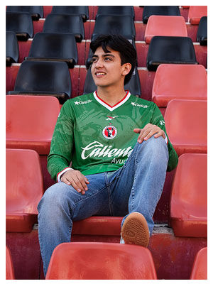 Gilberto Mora sentado en butacas rojas del estadio Caliente vistiendo jersey verde manga larga Edici&oacute;n Especial Reebok x Xoloitzcuintles, mano sobre rodilla, jeans azules, fondo de gradas rojas y negras vac&iacute;as.