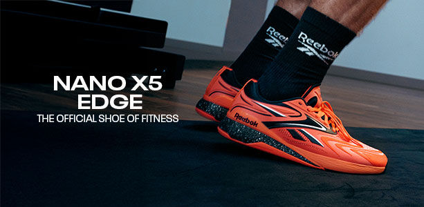 Banner desktop secci&oacute;n Reebok Nano X5 Edge, par de tenis crossfit naranja ne&oacute;n con detalles negros y plateados sobre colchoneta de gimnasio, calcetines negros con logo Reebok, texto Nano X5 Edge The Official Shoe of Fitness, fondo de piso de madera y equipo de entrenamiento oscuro.
