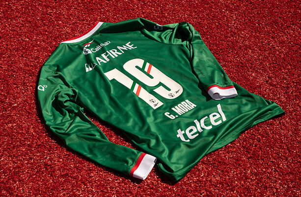 Vista plana trasera del jersey verde manga larga Reebok x Club Tijuana sobre c&eacute;sped rojo, n&uacute;mero 19 con franjas tricolor, nombre G. MORA, logo Reebok, pu&ntilde;os en blanco y rojo.