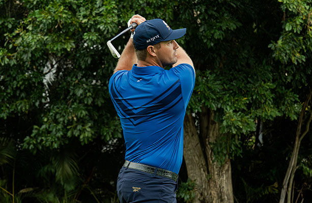 Modelo Reebok Golf de espaldas en polo azul con detalles oscuros, gorra azul, swing con hierro, fondo de &aacute;rboles y vegetaci&oacute;n.