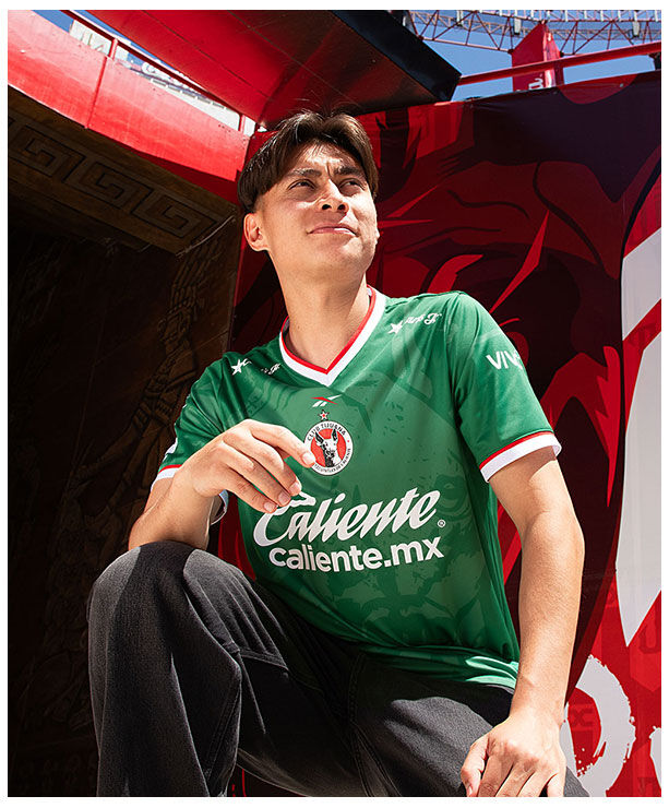 Card Reebok x Xolos con modelo en jersey verde sentado, mural rojo de fondo.