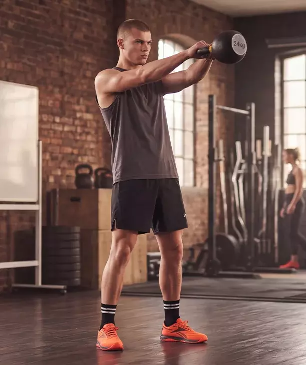 Look mobile de entrenamiento Reebok para hombre, modelo realizando kettlebell swing con pesa rusa de 24 kg en gimnasio de ladrillo, tank top gris, short negro Reebok, calcetines negros con rayas blancas y tenis de training naranjas Reebok Nano, rack de pesas al fondo. Ropa deportiva Reebok para hombre, tenis de training naranjas, outfit de gimnasio y crossfit.