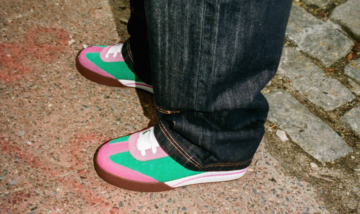 Imagen carrusel Reebok x IceCream Board Flip, modelo con jeans oscuros calzando tenis skate en combinaci&oacute;n verde rosa y blanco con suela caf&eacute;, sobre pavimento rojizo con adoquines grises, dise&ntilde;o retro con costuras contrastantes.