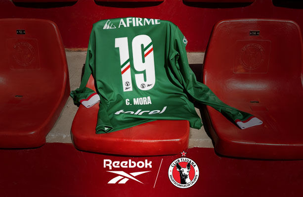 Jersey verde manga larga n&uacute;mero 19 G. MORA colocado sobre butacas rojas del estadio Caliente, logos Reebok y Club Tijuana Xoloitzcuintles en parte inferior, colecci&oacute;n Drop 4 Edici&oacute;n Especial.