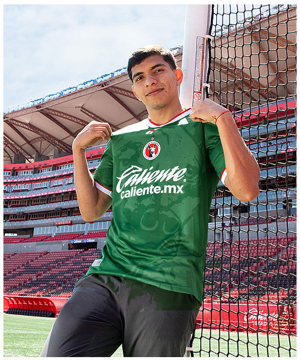 Modelo masculino sosteniendo el cuello del jersey verde manga corta Reebok x Club Tijuana frente a la porter&iacute;a del estadio Caliente, escudo Xoloitzcuintles y logo Reebok visibles, fondo de butacas rojas y estructura met&aacute;lica.