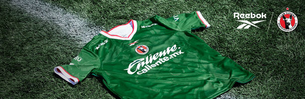 Banner horizontal con jersey verde manga corta Reebok x Club Tijuana extendido sobre c&eacute;sped del estadio Caliente, escudo Xoloitzcuintles, logo Reebok, branding Reebok y Club Tijuana en esquina superior derecha.