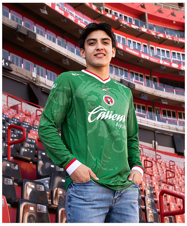 Gilberto Mora posando en gradas del estadio Caliente con jersey verde manga larga Edici&oacute;n Especial Reebok x Club Tijuana, escudo Xoloitzcuintles en pecho, logo Reebok visible, fondo de butacas rojas y negras del estadio, colecci&oacute;n oficial Reebok.