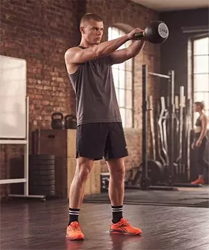 Look mobile de entrenamiento Reebok para hombre, modelo realizando kettlebell swing con pesa rusa de 24 kg en gimnasio de ladrillo, tank top gris, short negro Reebok, calcetines negros con rayas blancas y tenis de training naranjas Reebok Nano, rack de pesas al fondo. Ropa deportiva Reebok para hombre, tenis de training naranjas, outfit de gimnasio y crossfit.