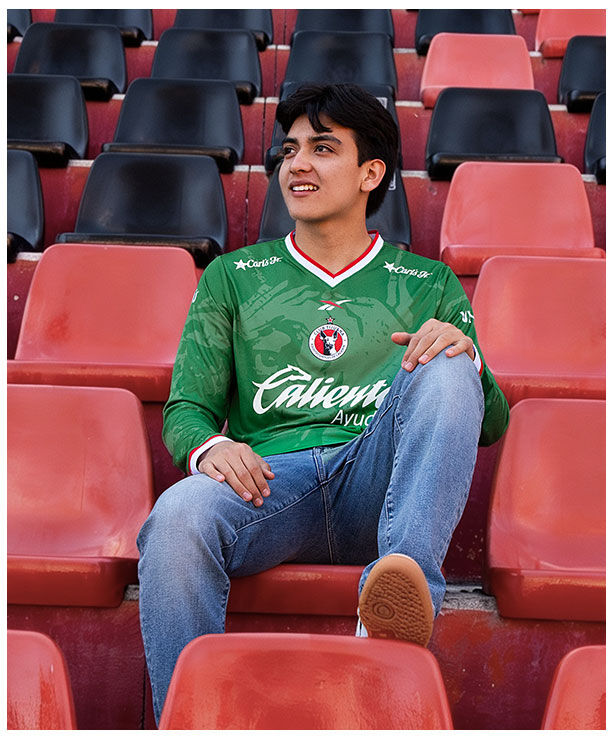 Gilberto Mora sentado en butacas rojas del estadio Caliente vistiendo jersey verde manga larga Edici&oacute;n Especial Reebok x Xoloitzcuintles, mano sobre rodilla, jeans azules, fondo de gradas rojas y negras vac&iacute;as.