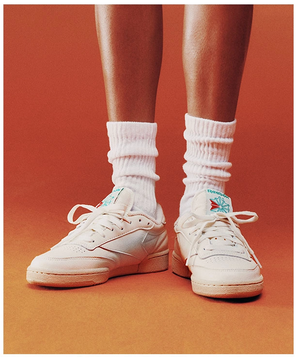 Portrait Reebok x Karol G Born Classic con primer plano de piernas, calcetines blancos altos y tenis Reebok Club C 85 blancos con logo Starcrest verde y rojo sobre fondo naranja. Reebok, Karol G, Club C 85, tenis blancos Reebok, Reebok Classics y campa&ntilde;a Born Classic Worn For Life