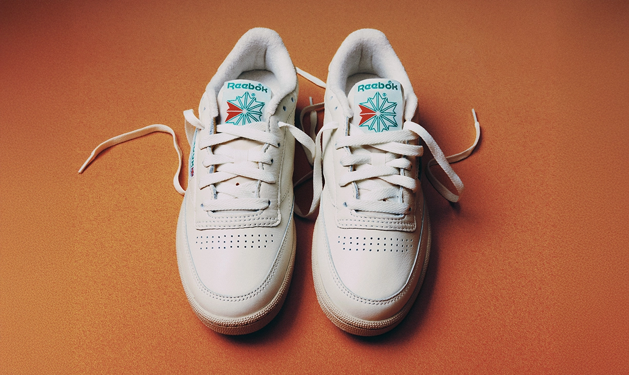 Par de tenis Reebok Club C 85 blancos vistos desde arriba sobre fondo naranja texturizado, con logo Reebok Starcrest verde y rojo en la leng&uuml;eta y agujetas sueltas, campa&ntilde;a Reebok x Karol G Born Classic. Reebok, Karol G, Club C 85, tenis blancos Reebok, Reebok Classics y Born Classic Worn For Life.