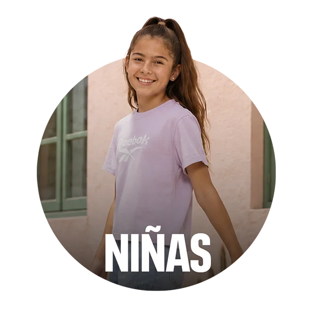 Card circular de categor&iacute;a Ni&ntilde;as Reebok, ni&ntilde;a sonriente de cabello largo con playera lila de logo Reebok en tono a tono, fondo de fachada rosa con ventanas verdes, texto Ni&ntilde;as en blanco. Ropa infantil de ni&ntilde;a Reebok .