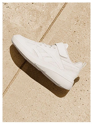 Portrait Reebok Back to School, tenis blanco Reebok con correa de velcro y upper textil, vista de perfil sobre piso de baldosas beige con luz natural. Tenis Reebok blancos, tenis blancos para ni&ntilde;o, tenis blancos con velcro y tenis para la escuela