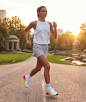 Look de running Reebok SoleMate para mujer, modelo corriendo en carretera al atardecer, tank top blanco Reebok, short gris, calcetines blancos y tenis de running blancos Reebok con suela multicolor arco&iacute;ris, parque y edificios al fondo con luz c&aacute;lida. Ropa deportiva Reebok para mujer, tenis de running blancos SoleMate y outfit para correr.