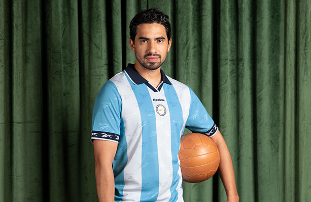 Modelo Reebok Jerseys Retro con jersey celeste y blanco a rayas, escudo retro, bal&oacute;n vintage, fondo cortina verde.