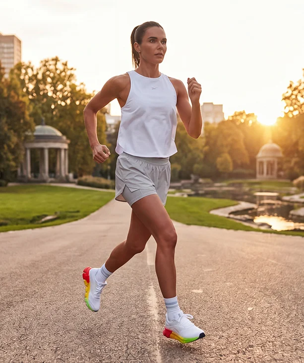 Look de running Reebok SoleMate para mujer, modelo corriendo en carretera al atardecer, tank top blanco Reebok, short gris, calcetines blancos y tenis de running blancos Reebok con suela multicolor arco&iacute;ris, parque y edificios al fondo con luz c&aacute;lida. Ropa deportiva Reebok para mujer, tenis de running blancos SoleMate y outfit para correr.