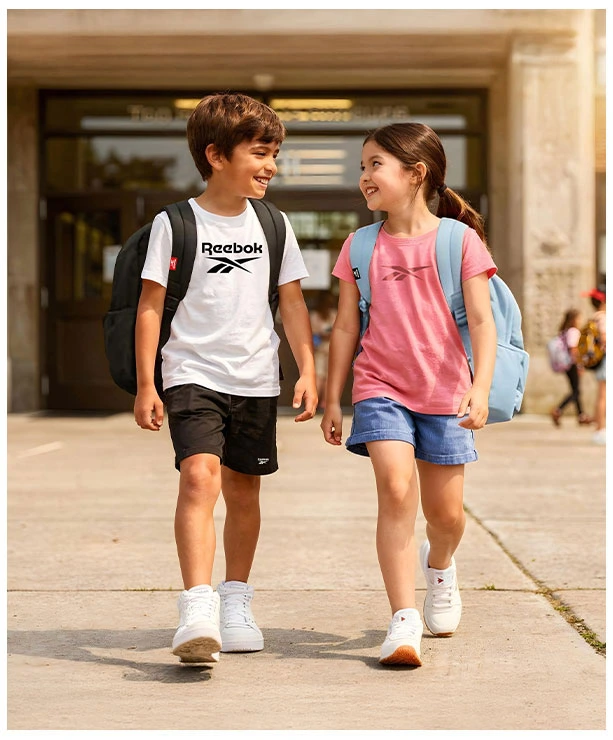 Portrait Reebok Back to School, ni&ntilde;o con playera blanca Reebok, short negro, mochila negra y tenis blancos caminando junto a ni&ntilde;a con playera rosa Reebok, short de mezclilla, mochila azul claro y tenis blancos, entrada de la escuela de fondo. Tenis Reebok blancos, tenis blancos para ni&ntilde;o y para ni&ntilde;a, tenis para la escuela.