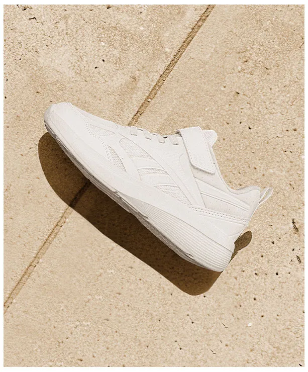 Portrait Reebok Back to School, tenis blanco Reebok con correa de velcro y upper textil, vista de perfil sobre piso de baldosas beige con luz natural. Tenis Reebok blancos, tenis blancos para ni&ntilde;o, tenis blancos con velcro y tenis para la escuela