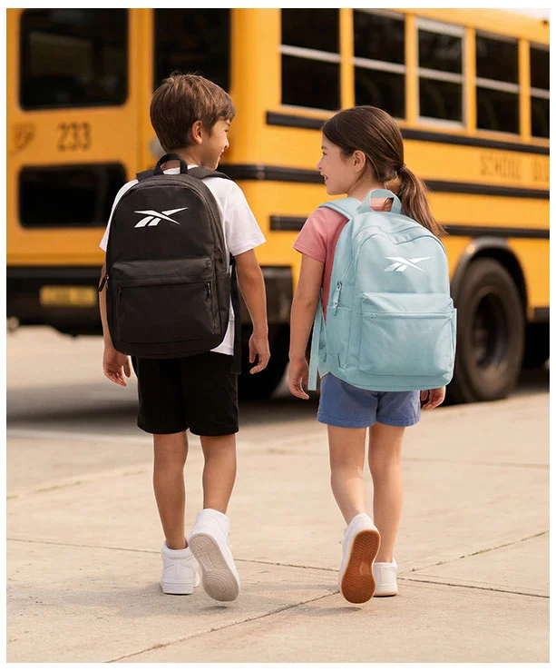 Portrait Reebok Back to School, ni&ntilde;o con mochila negra Reebok y ni&ntilde;a con mochila azul claro Reebok de espaldas caminando hacia autob&uacute;s escolar amarillo, ambos con tenis blancos. Mochilas Reebok, tenis blancos para ni&ntilde;os, tenis para la escuela y regreso a clases