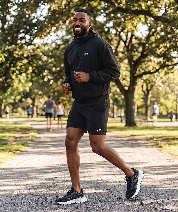 Look mobile de hombre Reebok, modelo corriendo en un parque con rompevientos negro con logo Reebok Vector, short negro y tenis de running negros con suela blanca. Ropa deportiva de hombre Reebok.