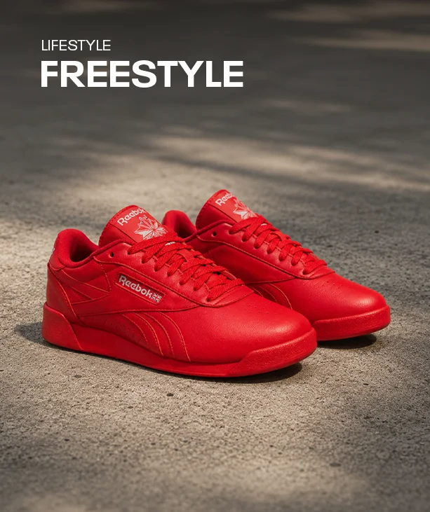 Silueta Reebok Freestyle de lifestyle, par de tenis rojos de piel de ca&ntilde;a baja con logo Reebok Vector, sobre piso de concreto con luz suave. Tenis lifestyle Freestyle Reebok.
