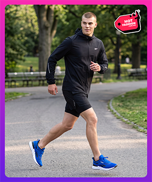 Look Reebok Hot Fashion para hombre, modelo corriendo por un sendero de parque arbolado con chamarra rompevientos negra Reebok con capucha y logo Vector, short negro y tenis de running azules Reebok con suela blanca, bancas y &aacute;rboles de fondo, etiqueta Hot Fashion. Reebok hombre, ropa deportiva Reebok, chamarra de running, tenis azules Reebok y rebajas Hot Fashion.