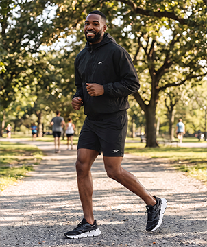 Look mobile de hombre Reebok, modelo corriendo en un parque con rompevientos negro con logo Reebok Vector, short negro y tenis de running negros con suela blanca. Ropa deportiva de hombre Reebok.