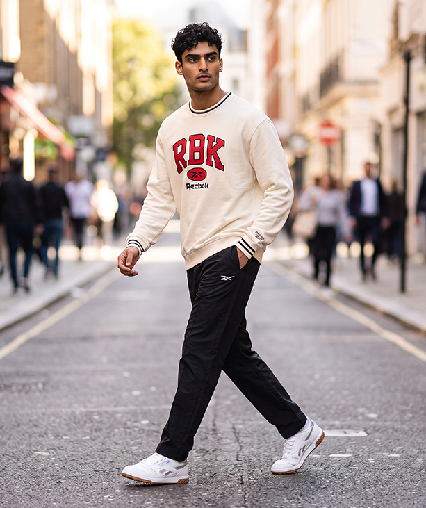 Look mobile de hombre Reebok, modelo caminando por una calle urbana con sudadera crema con letras RBK en rojo, pants negros con logo Reebok Vector y tenis Classic Leather blancos. Ropa de hombre Reebok
