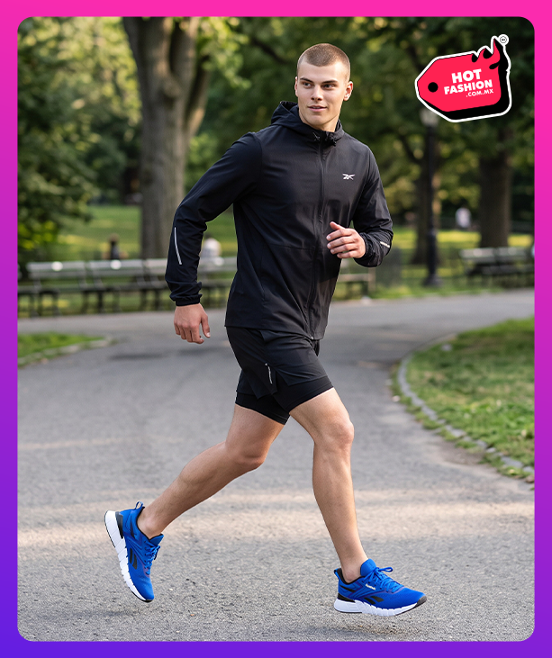 Look Reebok Hot Fashion para hombre, modelo corriendo por un sendero de parque arbolado con chamarra rompevientos negra Reebok con capucha y logo Vector, short negro y tenis de running azules Reebok con suela blanca, bancas y &aacute;rboles de fondo, etiqueta Hot Fashion. Reebok hombre, ropa deportiva Reebok, chamarra de running, tenis azules Reebok y rebajas Hot Fashion.