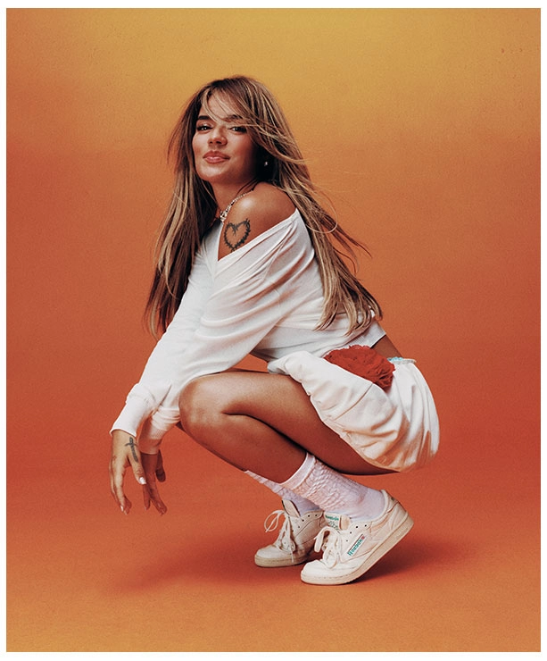 Portrait Reebok x Karol G Born Classic, Karol G en cuclillas con conjunto blanco oversize de hombros descubiertos, tatuaje de coraz&oacute;n, calcetines blancos y tenis Reebok Club C 85 blancos sobre fondo naranja. Reebok, Karol G, Club C 85, tenis blancos Reebok, Reebok Classics y campa&ntilde;a Born Classic Worn For Life.