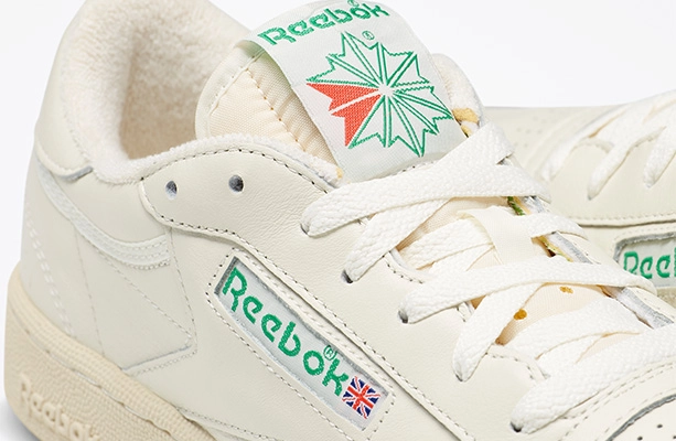 Detalle de los tenis Reebok Club C 85 en color crema mostrando la leng&uuml;eta con logo Reebok Starcrest verde y rojo, el logo Union Jack en el costado y la piel perforada, campa&ntilde;a Reebok x Karol G Born Classic. Reebok, Karol G, Club C 85, tenis blancos Reebok, Reebok Classics y Born Classic Worn For Life.