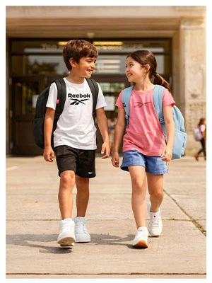 Portrait Reebok Back to School, ni&ntilde;o con playera blanca Reebok, short negro, mochila negra y tenis blancos caminando junto a ni&ntilde;a con playera rosa Reebok, short de mezclilla, mochila azul claro y tenis blancos, entrada de la escuela de fondo. Tenis Reebok blancos, tenis blancos para ni&ntilde;o y para ni&ntilde;a, tenis para la escuela.