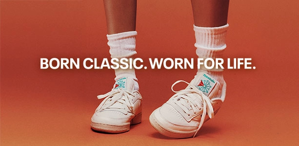 Banner secundario desktop Reebok x Karol G con texto Born Classic Worn For Life, piernas con calcetines blancos altos y tenis Reebok Club C 85 blancos con detalles verde y rojo sobre fondo naranja. Reebok, Karol G, Club C 85, tenis blancos Reebok, Reebok Classics y campa&ntilde;a Born Classic Worn For Life.