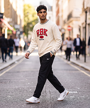 Look mobile de hombre Reebok, modelo caminando por una calle urbana con sudadera crema con letras RBK en rojo, pants negros con logo Reebok Vector y tenis Classic Leather blancos. Ropa de hombre Reebok