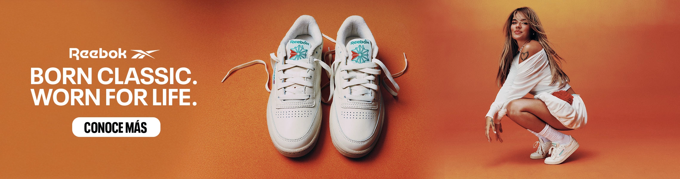 Banner desktop home Reebok x Karol G Born Classic Worn For Life, Karol G en cuclillas con conjunto blanco oversize y tenis Club C 85 blancos, par de tenis Club C 85 en primer plano sobre fondo naranja, logo Reebok Vector y bot&oacute;n Conoce M&aacute;s. Reebok, Karol G, Club C 85, tenis blancos Reebok, Reebok Classics y campa&ntilde;a Born Classic Worn For Life.
