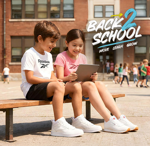 Head banner desktop Reebok Back to School, ni&ntilde;o con playera blanca Reebok y ni&ntilde;a con playera rosa viendo una tablet en banca frente al edificio escolar con estudiantes de fondo, texto Back 2 School Move Learn Grow. Tenis Reebok blancos, tenis blancos para ni&ntilde;os, tenis para la escuela y regreso a clases.