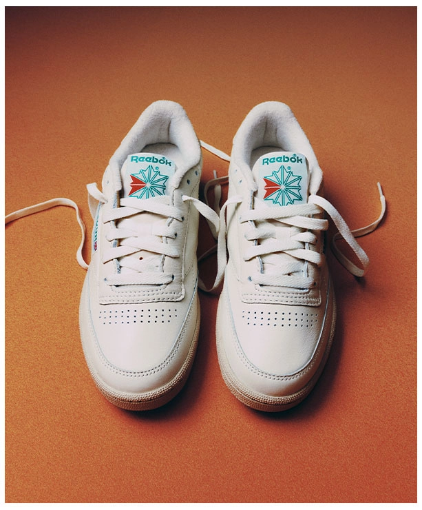 Portrait Reebok x Karol G Born Classic con par de tenis Reebok Club C 85 blancos vistos desde arriba sobre fondo naranja, logo Reebok Starcrest verde y rojo en la leng&uuml;eta y agujetas sueltas. Reebok, Karol G, Club C 85, tenis blancos Reebok, Reebok Classics y campa&ntilde;a Born Classic Worn For Life.