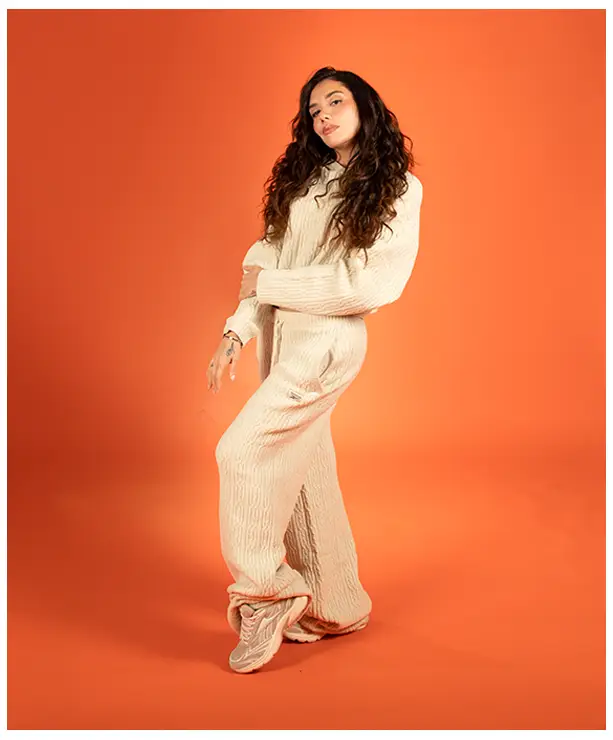 Imagen del carrusel de la landing Reebok Born Classic Worn Together FW26, modelo de pie sobre fondo naranja con conjunto tejido color crema de su&eacute;ter y pants amplios Reebok y tenis claros. Reebok, Born Classic Worn Together, ropa Reebok FW26, conjunto tejido Reebok y Reebok Classics.