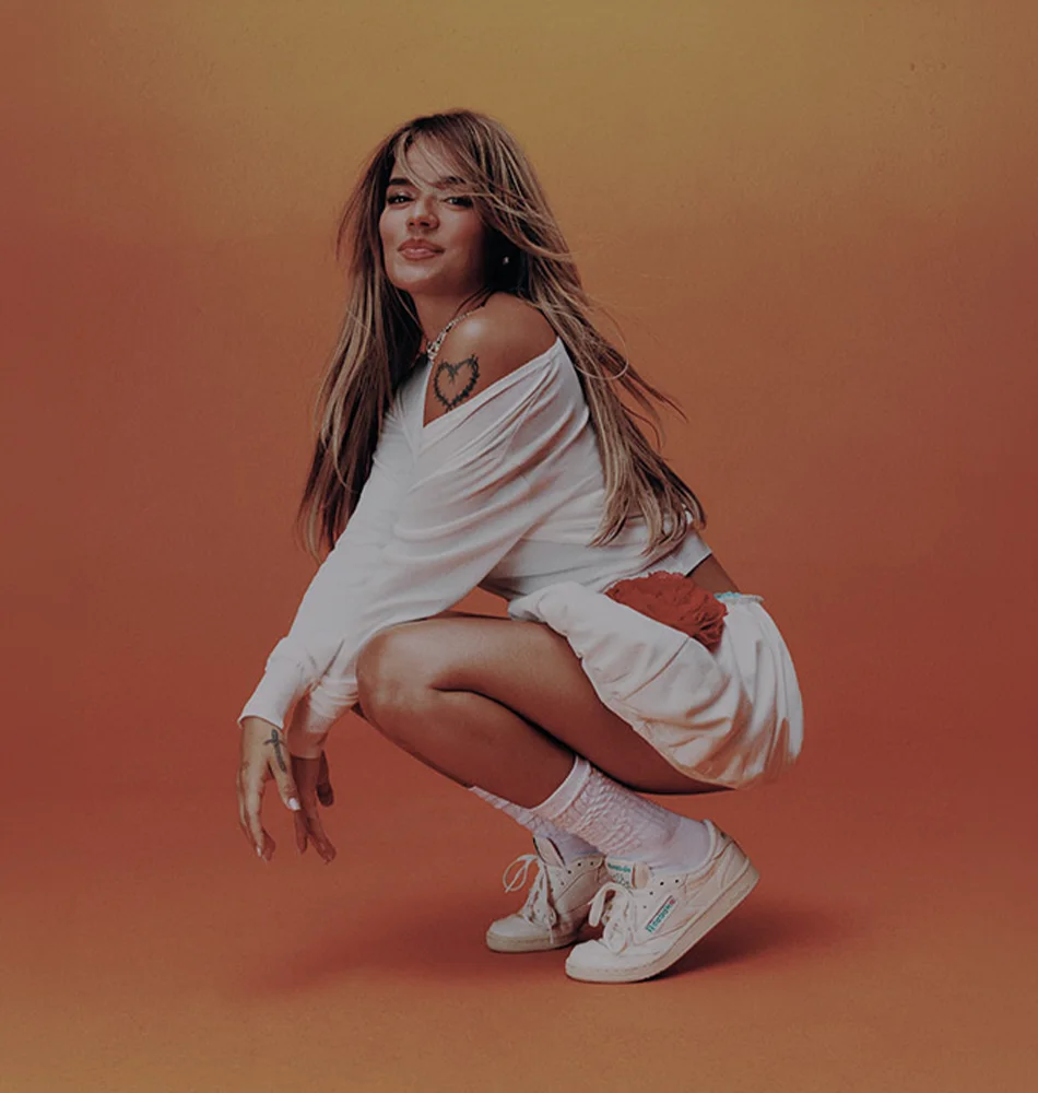 Karol G con outfit blanco y tenis Reebok en campa&ntilde;a sobre fondo naranja