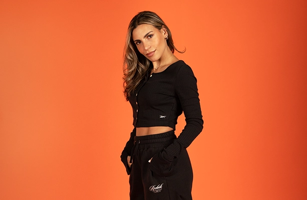 Imagen editorial de la landing Reebok Born Classic Worn Together FW26, modelo sobre fondo naranja con top negro de botones de manga larga y pants negros Reebok. Reebok, Born Classic Worn Together, ropa Reebok FW26, conjunto negro Reebok y Reebok Classics